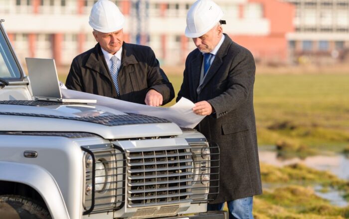 Two men in suits look at plans on the bonnet of a car while wearing hard hats