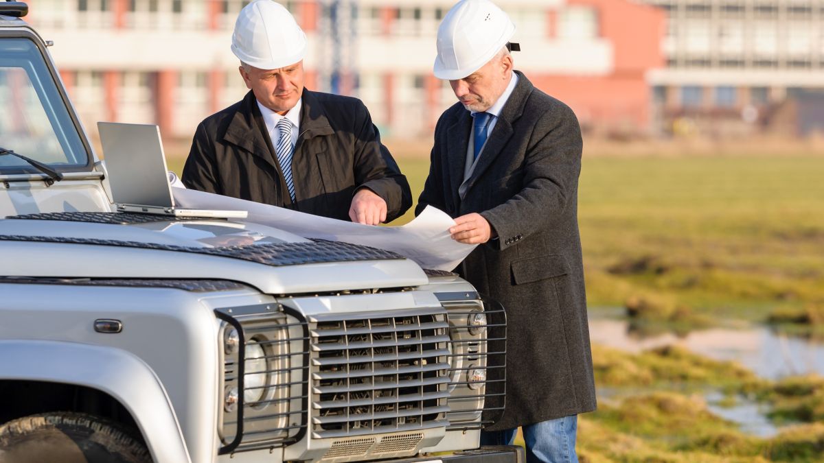 Two men in suits look at plans on the bonnet of a car while wearing hard hats